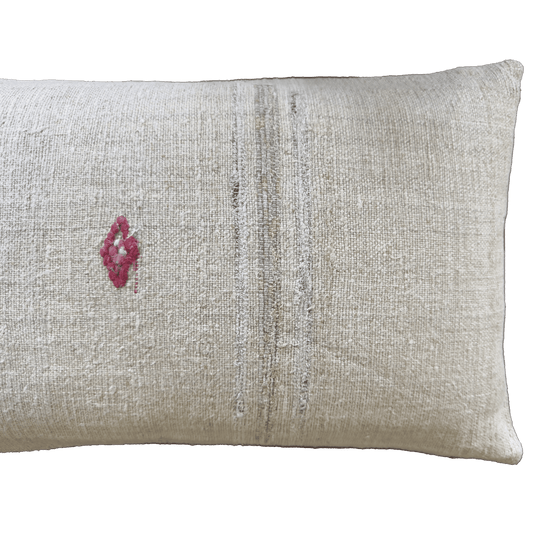 A rectangular beige linen throw pillow with a small pink embroidered diamond on the left and vertical distressed stripes in muted gray and olive tones.