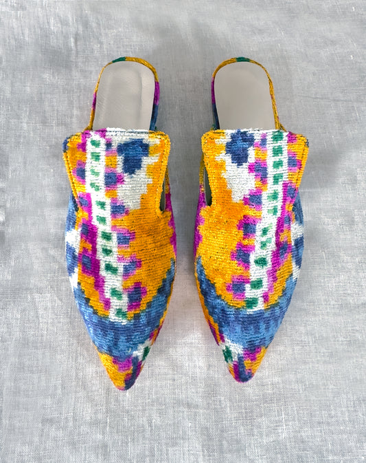 A pair of silk velvet ikat handmade slippers in orange, with sharp pointed toes, displayed on a white textured surface.