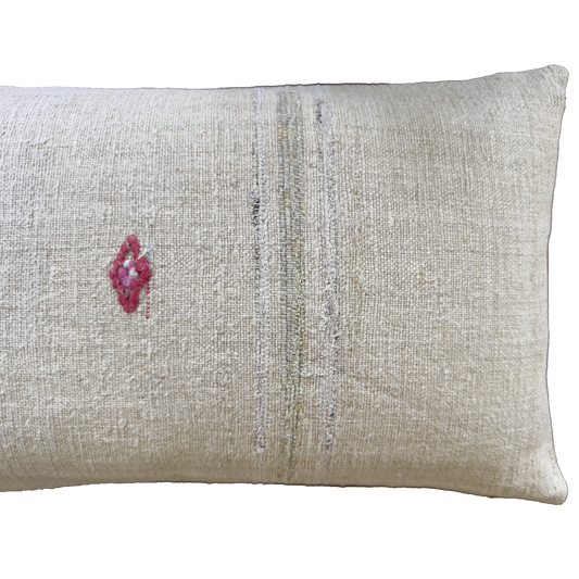 A rectangular beige linen throw pillow with a small pink embroidered diamond on the left and vertical distressed stripes in muted gray and olive tones.