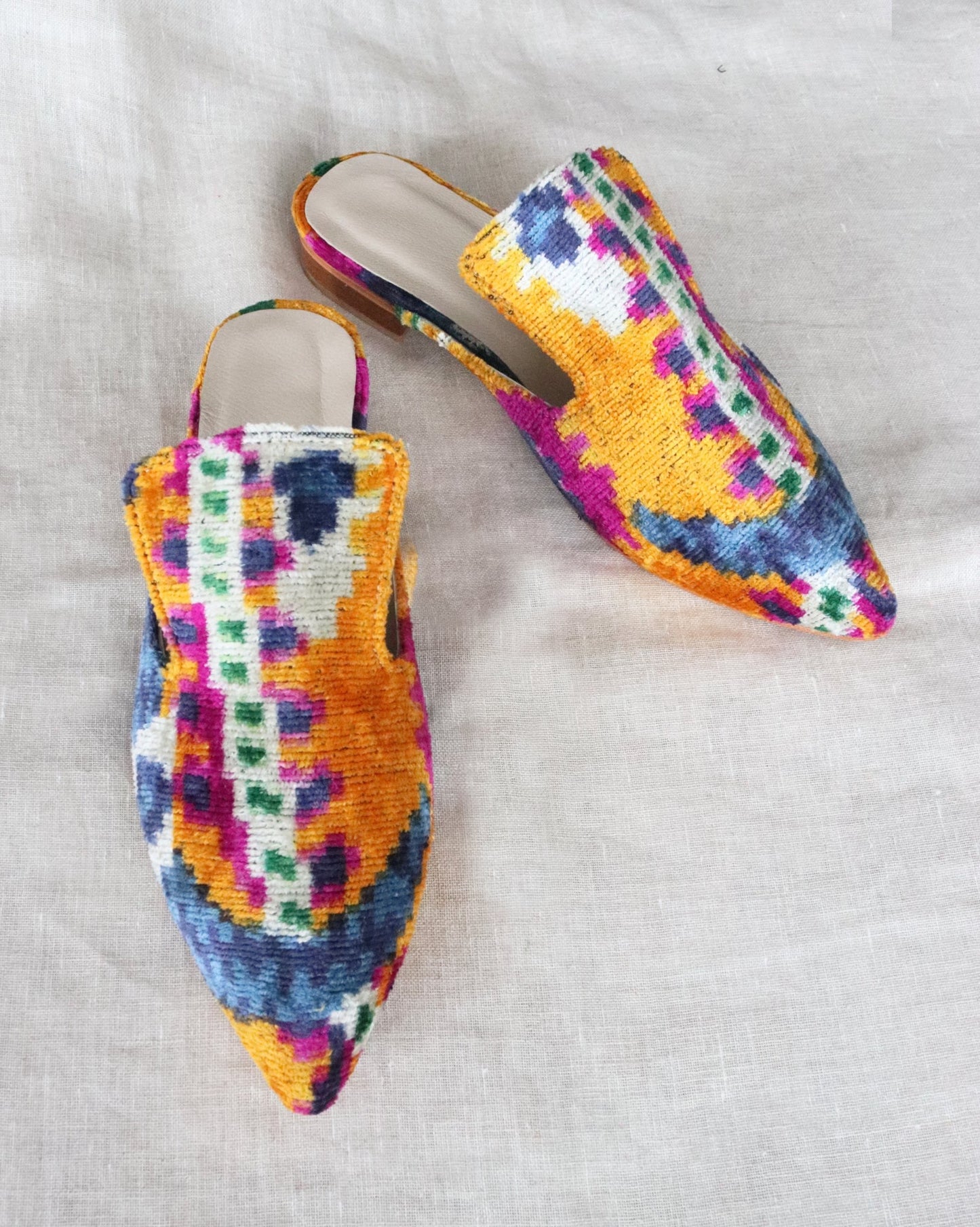A pair of silk velvet ikat handmade slippers in orange, with sharp pointed toes, displayed on a white textured surface.