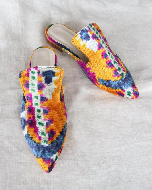 A pair of silk velvet ikat handmade slippers in orange, with sharp pointed toes, displayed on a white textured surface.