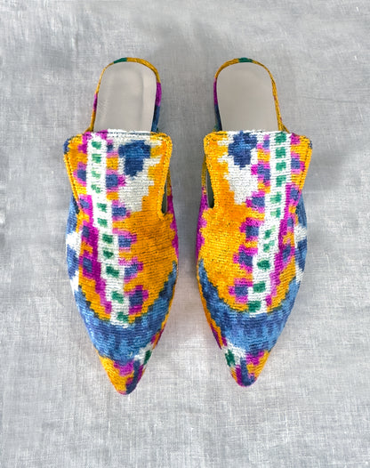 A pair of silk velvet ikat handmade slippers in orange, with sharp pointed toes, displayed on a white textured surface.