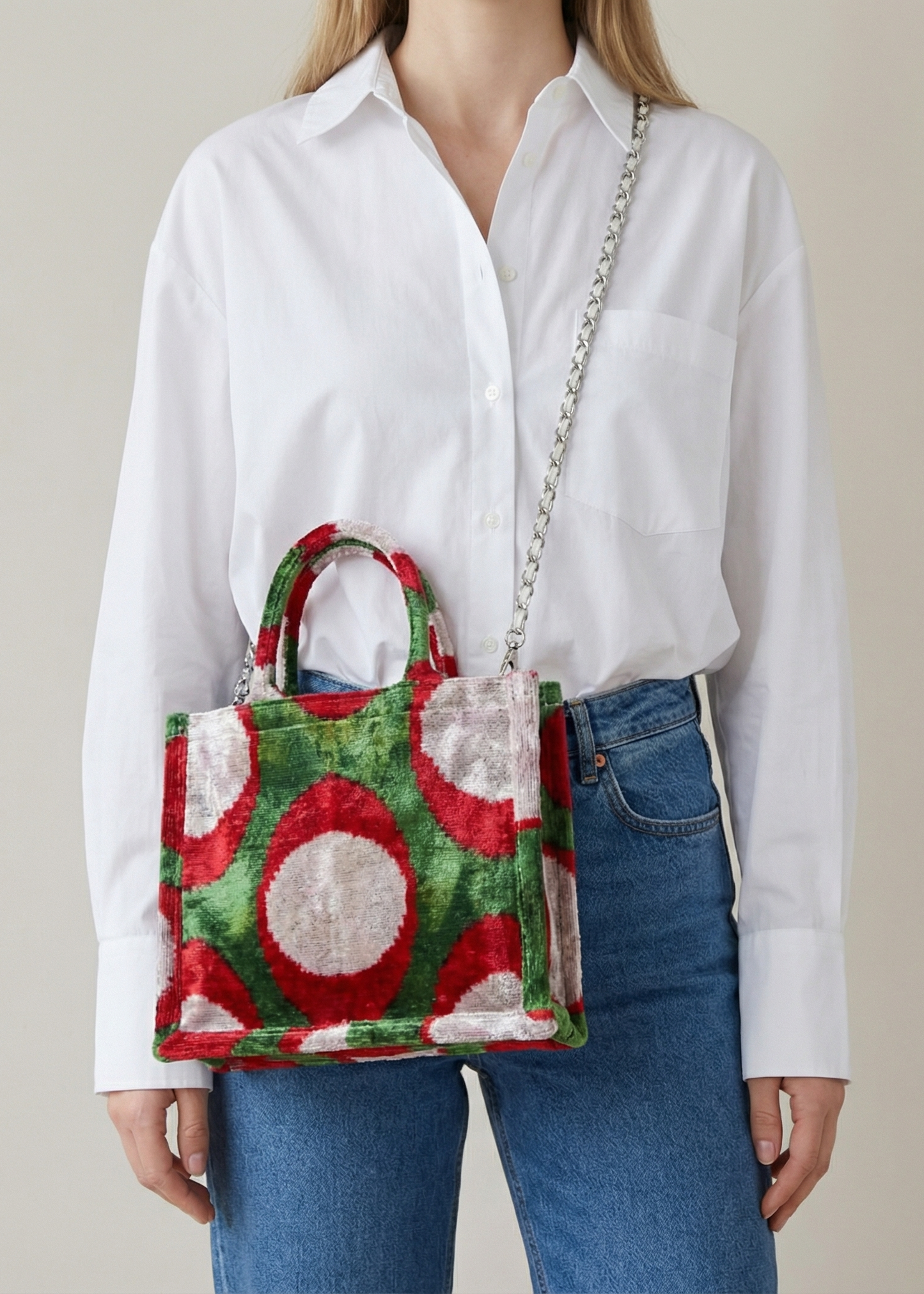Person wearing a white shirt and blue jeans holding a colorful handbag with a pattern of red and green shapes.
